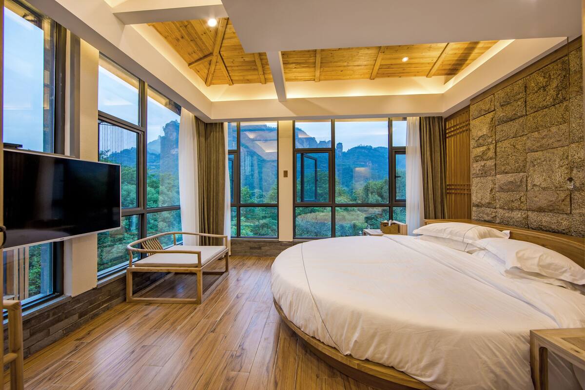 A bedroom with a large bed, a TV, and a view of a mountainous landscape through large windows.