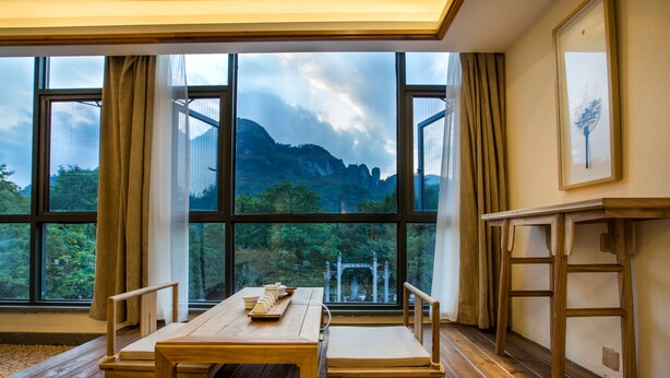 Una habitación con una mesa y sillas de madera, ventanas amplias con vistas a montañas y árboles, y un cuadro en la pared.