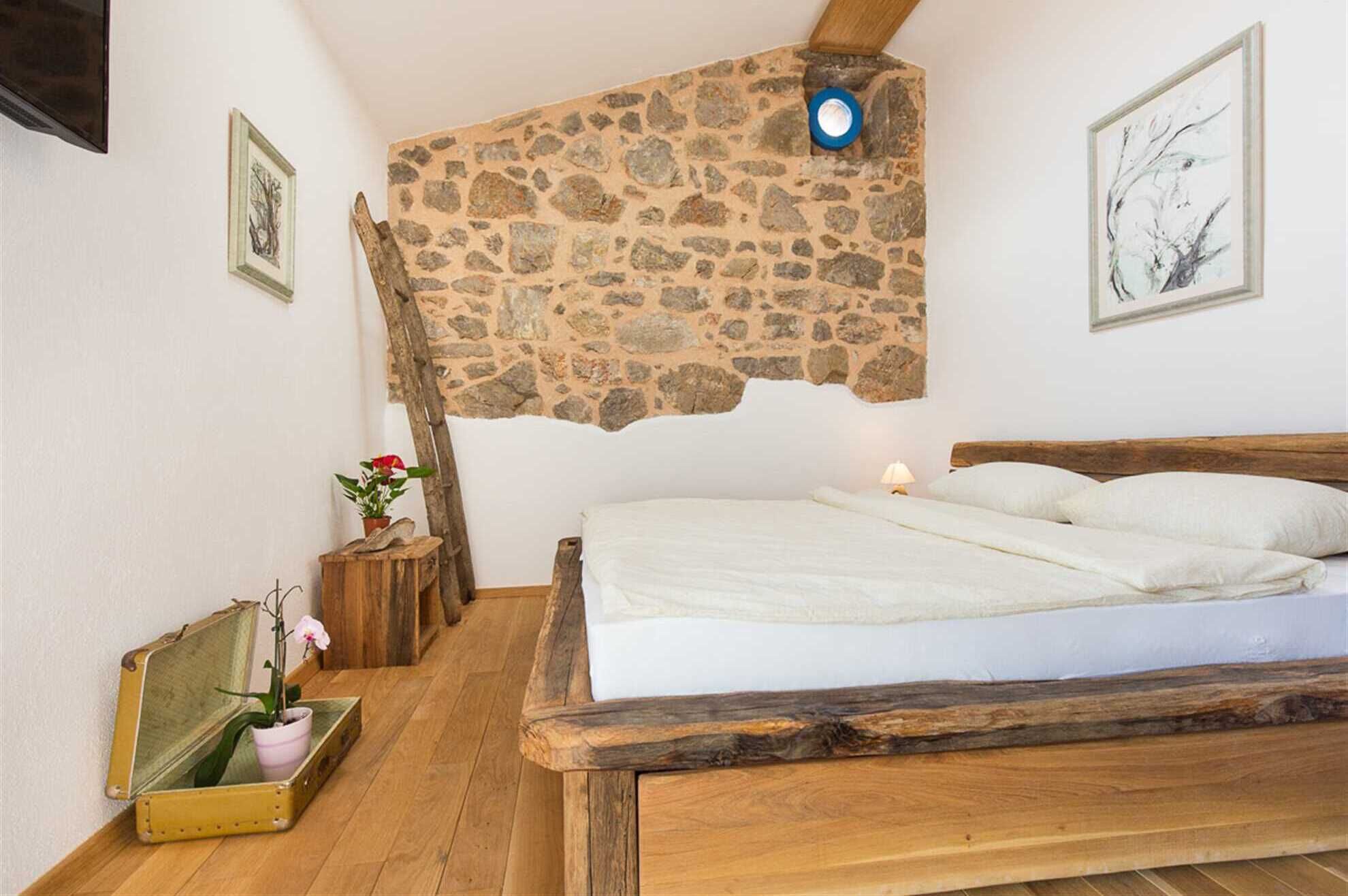 A bedroom with a stone wall, wooden bed frame, and a small bedside table with a lamp.