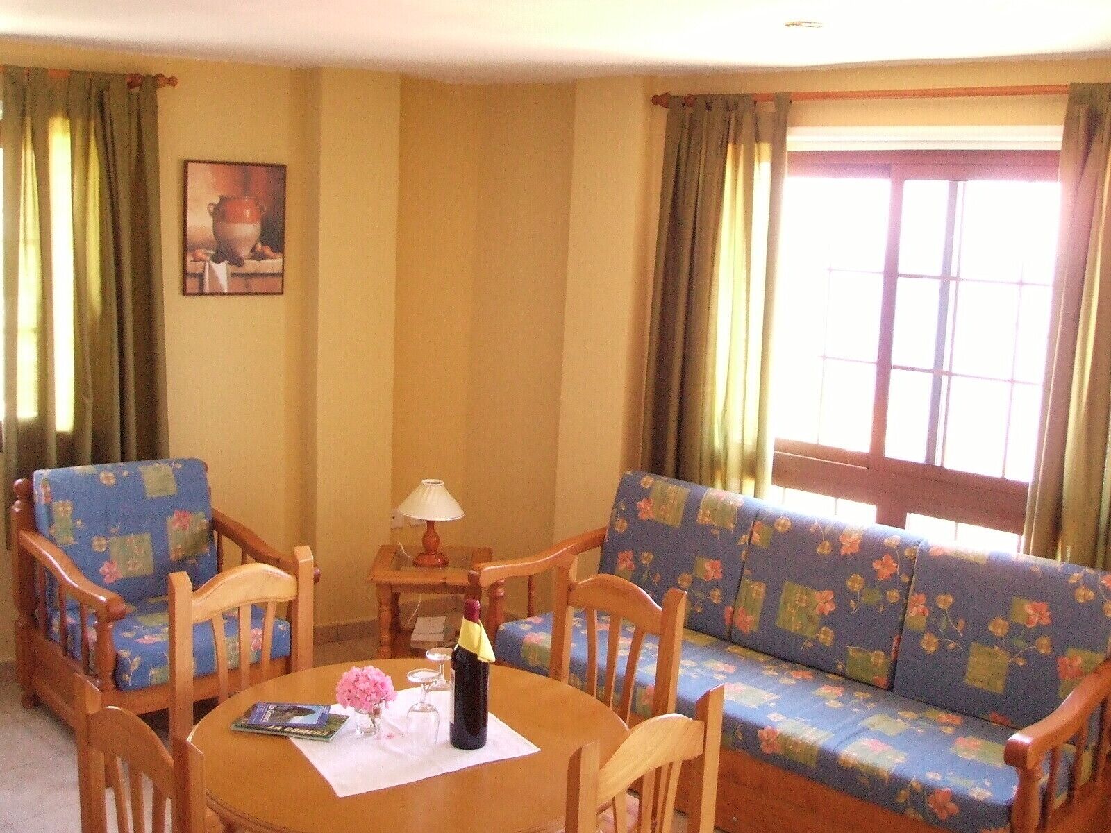 Uma sala de estar acolhedora com uma mesa redonda de madeira, sofá com padrão floral e cadeiras de madeira. Uma luminária está sobre a mesa, e há uma garrafa sobre a toalha.