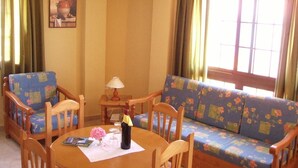 A cozy living room with a round wooden table, floral-patterned sofa, and wooden chairs. A lamp is on the table, and a bottle is on the tablecloth. - Quiet apartment with sea views (Agulo)
