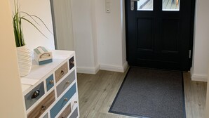 A clean, modern entryway with a black door, a white console table with drawers, a grey doormat, and a view of the outside balcony.