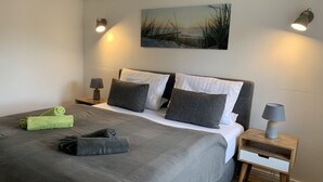 A neatly made bed with grey bedding, a green towel, and a grey blanket. There are two bedside tables with lamps, a painting on the wall, and a wooden floor.