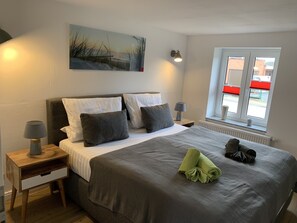 A neatly arranged bedroom with a large bed, two bedside tables, a window with blinds, and a painting on the wall.