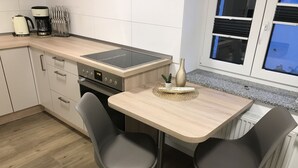 A modern kitchen with a built-in stove, two grey chairs, a small table with a decorative bowl, and a window with blinds.
