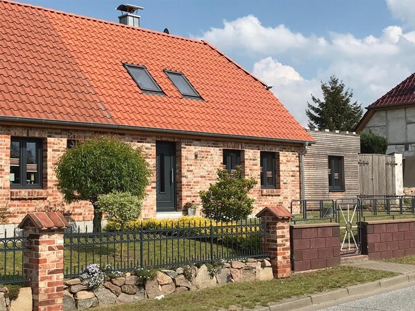 A two-story brick house with a red-tiled roof, two skylights, a front door, and a small front yard with a metal fence and a tree.
