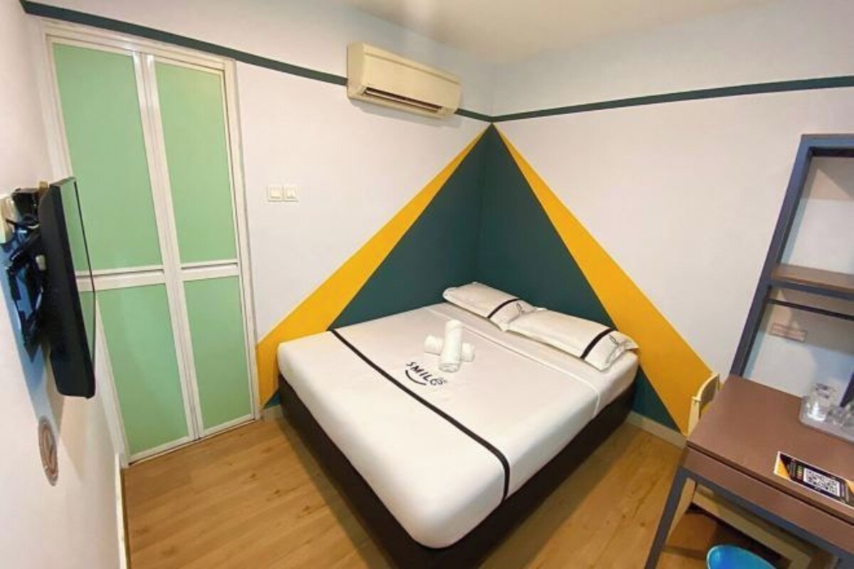 A small, compact room with a bed, a desk, and an air conditioning unit.