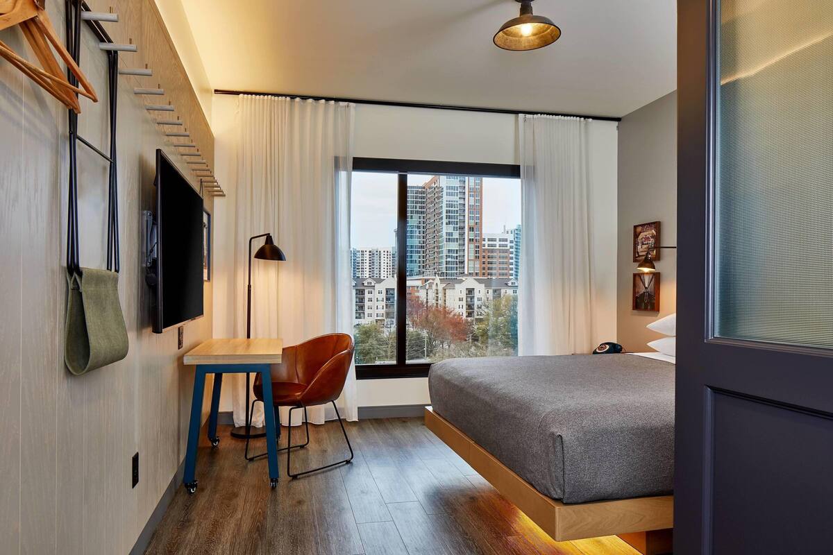 Una habitación de hotel moderna con cama, escritorio, silla, televisor y un amplio ventanal con vistas a la ciudad.