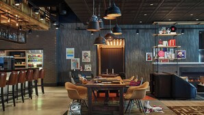 A modern bar area with a high ceiling, exposed wooden beams, and hanging pendant lights. There is a bar counter with stools and a refrigerator stocked with beverages. A fireplace is visible in the background, and a bookshelf with various items is on the right side. - Moxy Atlanta Midtown (Atlanta)