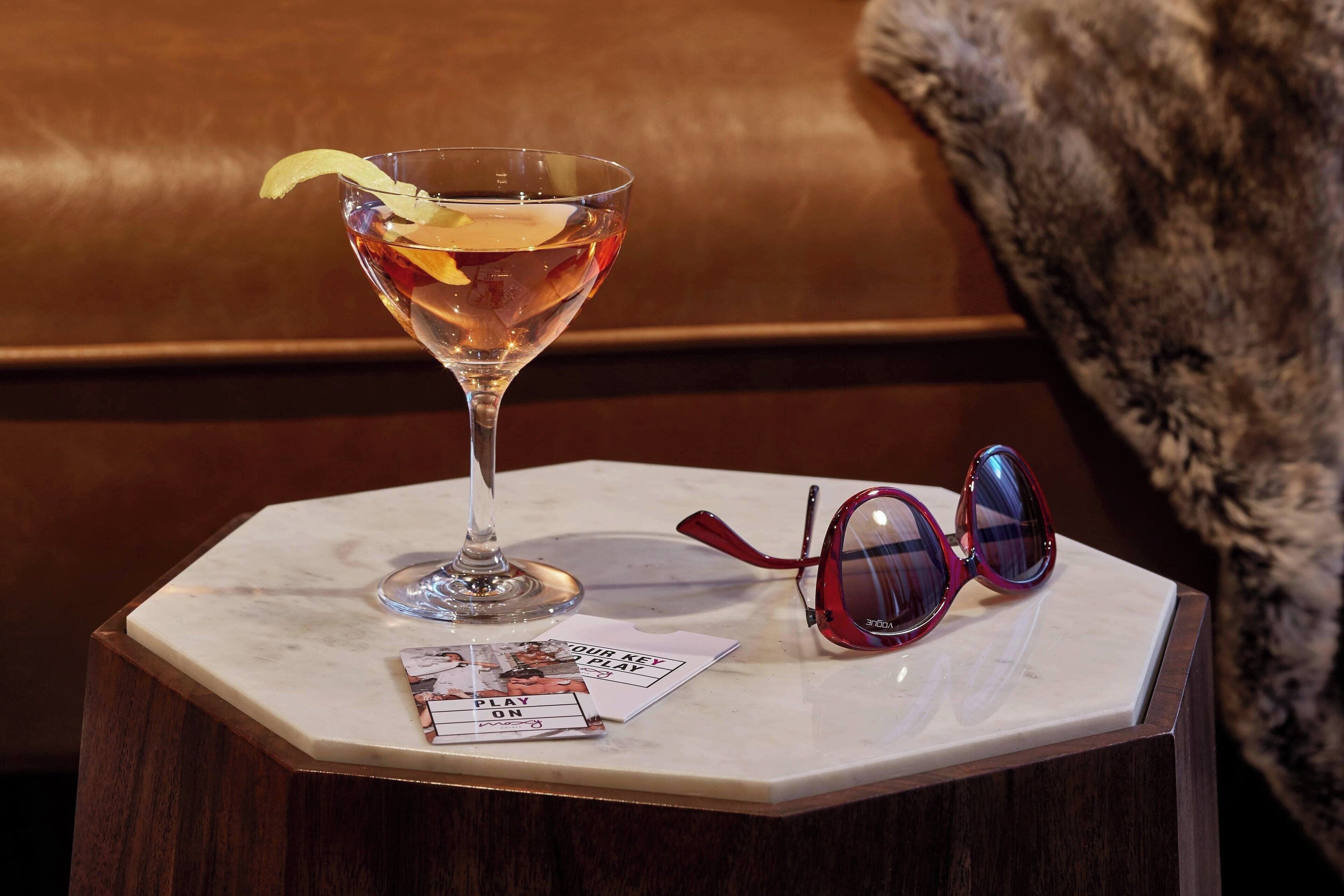 A cocktail glass with a lemon peel on a marble table, a pair of sunglasses, and a card with 'PLAY ON' text.
