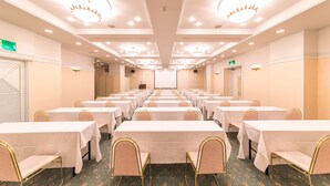 Un salón de conferencias con filas de mesas y sillas, equipado con luces en el techo y una pantalla para proyector.
