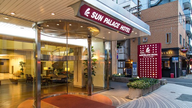 La entrada al Hotel Sun Palace con fachada de vidrio, un toldo rojo y una alfombra roja que conduce a la entrada.