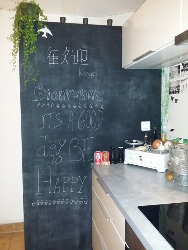 Une cuisine avec un mur recouvert d’un tableau noir où est écrit un message de bienvenue, et une plante suspendue au-dessus.
