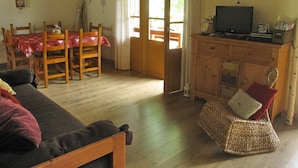 Een gezellige woonkamer met een bank, een eettafel met stoelen, een televisie op een houten kast, en een groot raam met gordijnen.