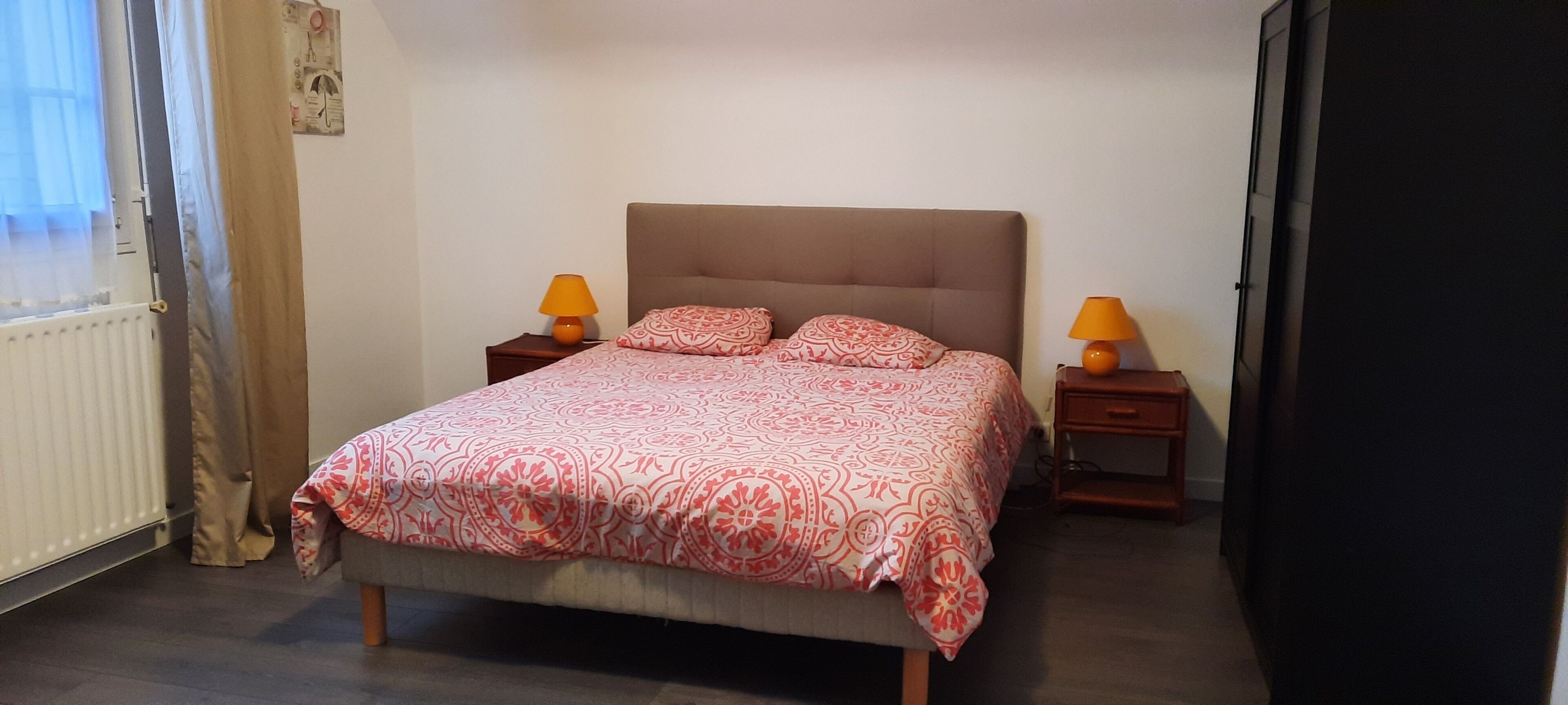 Une chambre à coucher avec un lit, deux tables de chevet, deux lampes, un radiateur et une fenêtre avec des rideaux.