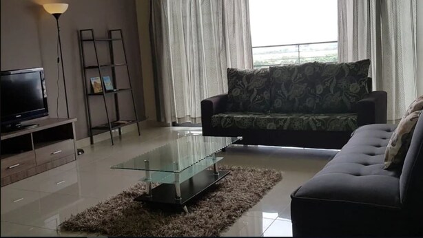 Una sala de estar con un sofá, una mesa de centro de vidrio, un televisor sobre un mueble bajo, una escalera y cortinas.
