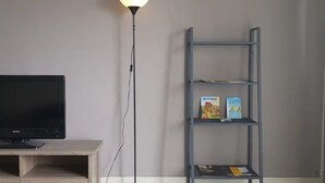 Una habitación con una televisión sobre un soporte de madera, una lámpara de pie y una estantería metálica llena de libros.