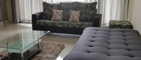 A living room with a glass coffee table, a tufted ottoman, a patterned sofa, and a patterned rug.