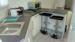 A compact kitchen with a sink, a microwave, a kettle, and a gas stove. There are two bar stools and a small window.