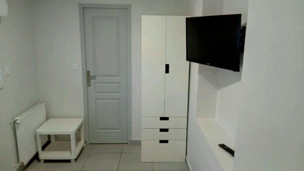 A small, white room with a door, a white cabinet with drawers, a small white table, and a mounted TV on the wall.