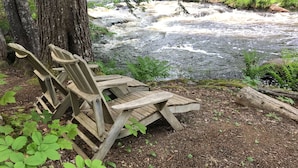 A wooden bench overlooking a river with a rocky bed, surrounded by greenery and trees. - A Quiet Riverside Cottage with 2 Fireplaces (Crousetown)