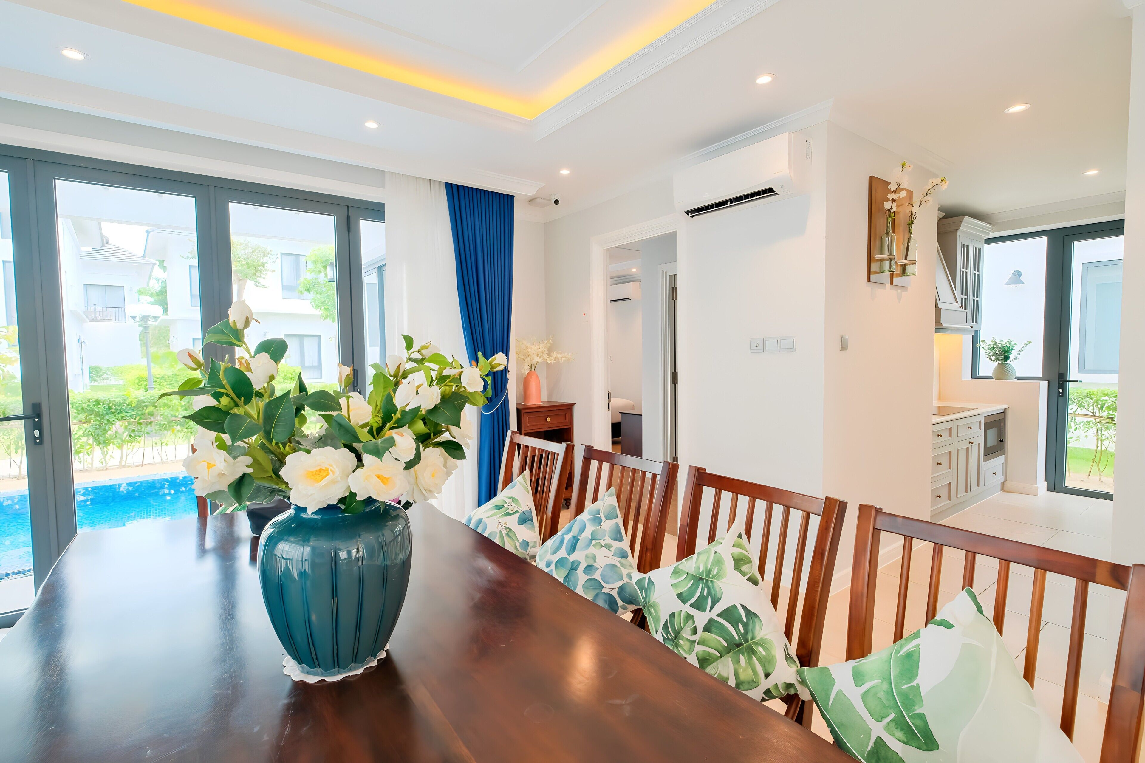 Un comedor con una mesa de madera, cojines blancos y azules con estampado floral, y un jarrón grande de flores.