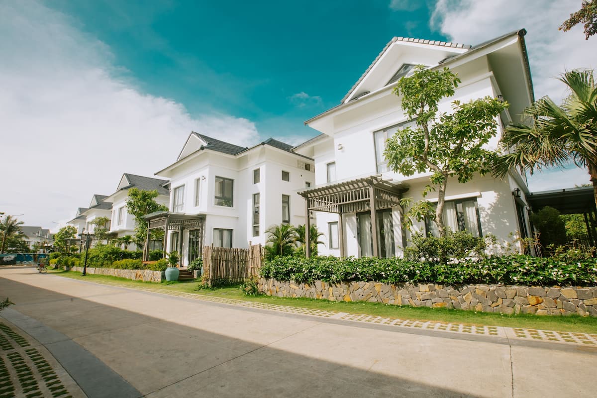 A row of modern white houses with stone walls and greenery, a clear sky, and a paved street.