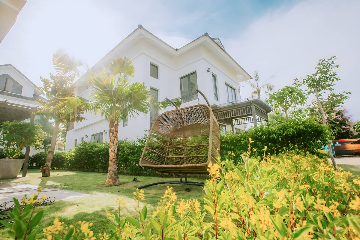 A modern house with a hanging chair in the garden, surrounded by lush greenery and palm trees.