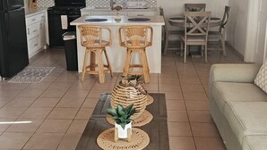 A spacious living area with a wooden ceiling, a kitchen with white cabinets, a dining area with wicker chairs, and a sofa.