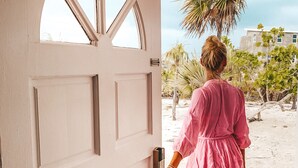 A woman in a pink dress exiting a white door onto a sandy beach with palm trees.