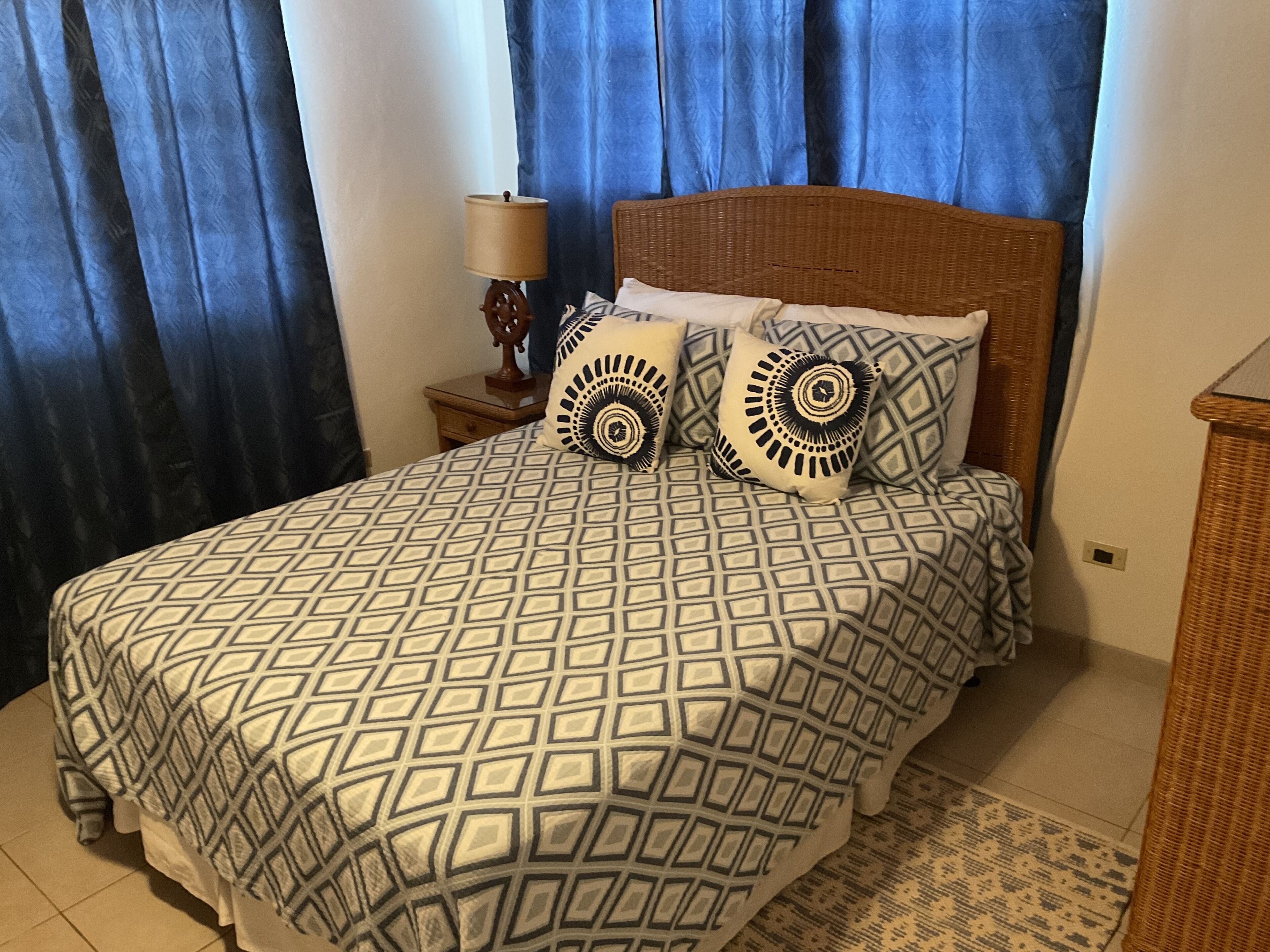 A bed with a geometric patterned bedspread, two pillows with circular designs, a wooden nightstand with a lamp, and blue curtains.