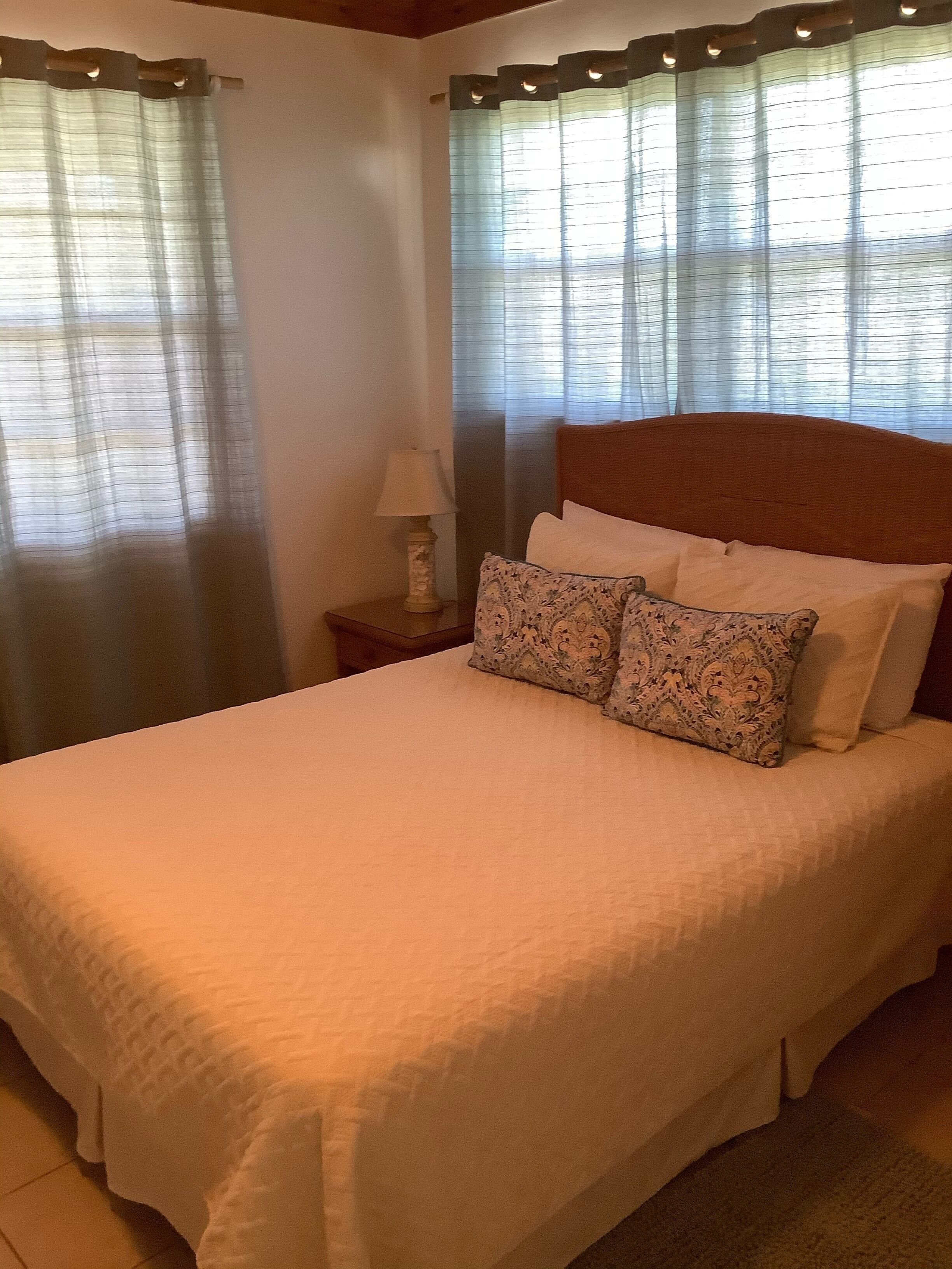 Una cama bien tendida con un almohadón estampado, una lámpara de noche y cortinas en una ventana.