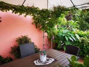 Een terras buiten met een houten tafel en stoelen, omringd door weelderige beplanting en een perzikkleurige muur.