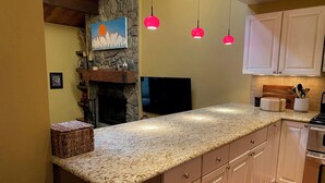 Una cocina con encimera de granito, armarios de madera, chimenea de piedra y lámparas colgantes.