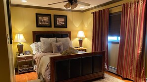 Un dormitorio con cama, dos mesitas de noche con lámparas, ventilador de techo y ventana con cortinas.