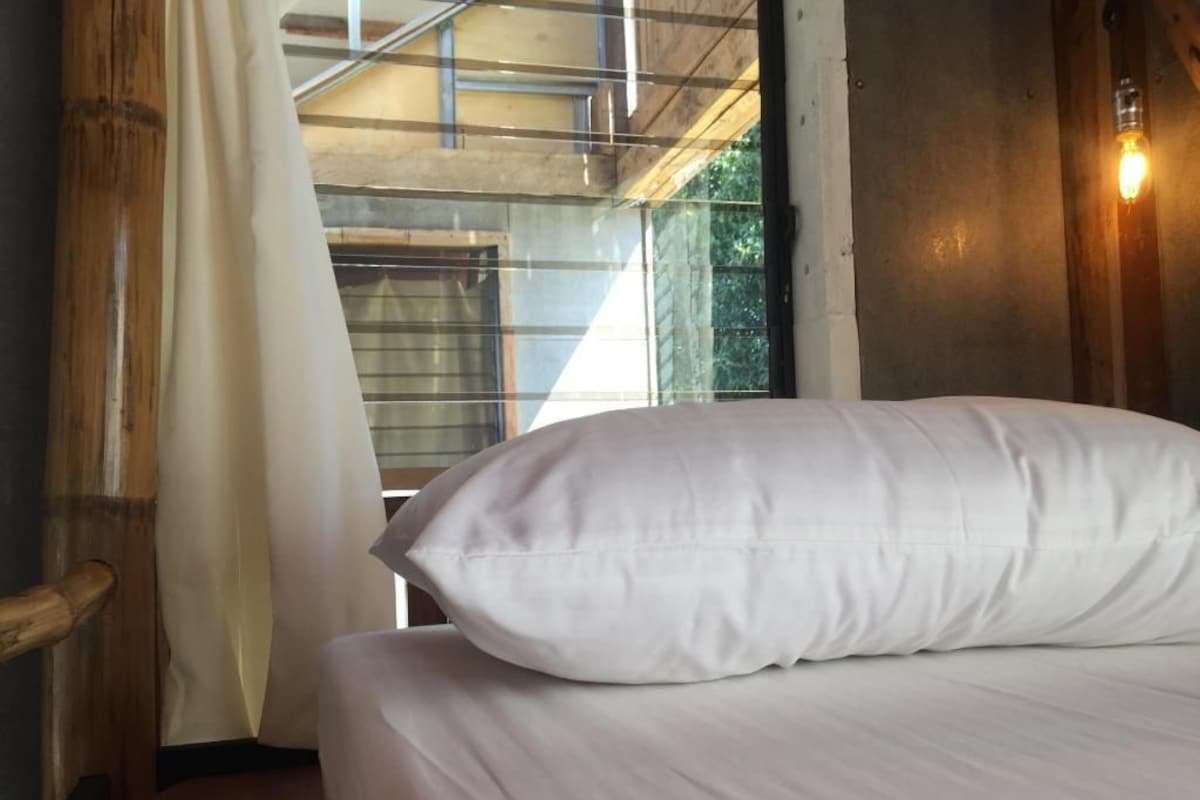 A bed with a white pillow in a room with a wooden floor, a window with a curtain, and a wall-mounted light.