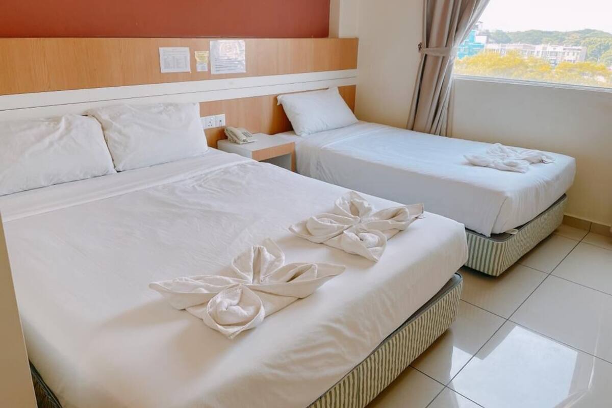 A hotel room with two beds, each with white linens and decorative towels. There is a nightstand with a phone and a wall-mounted clock. The room has a window with curtains, and the walls are painted in warm tones.