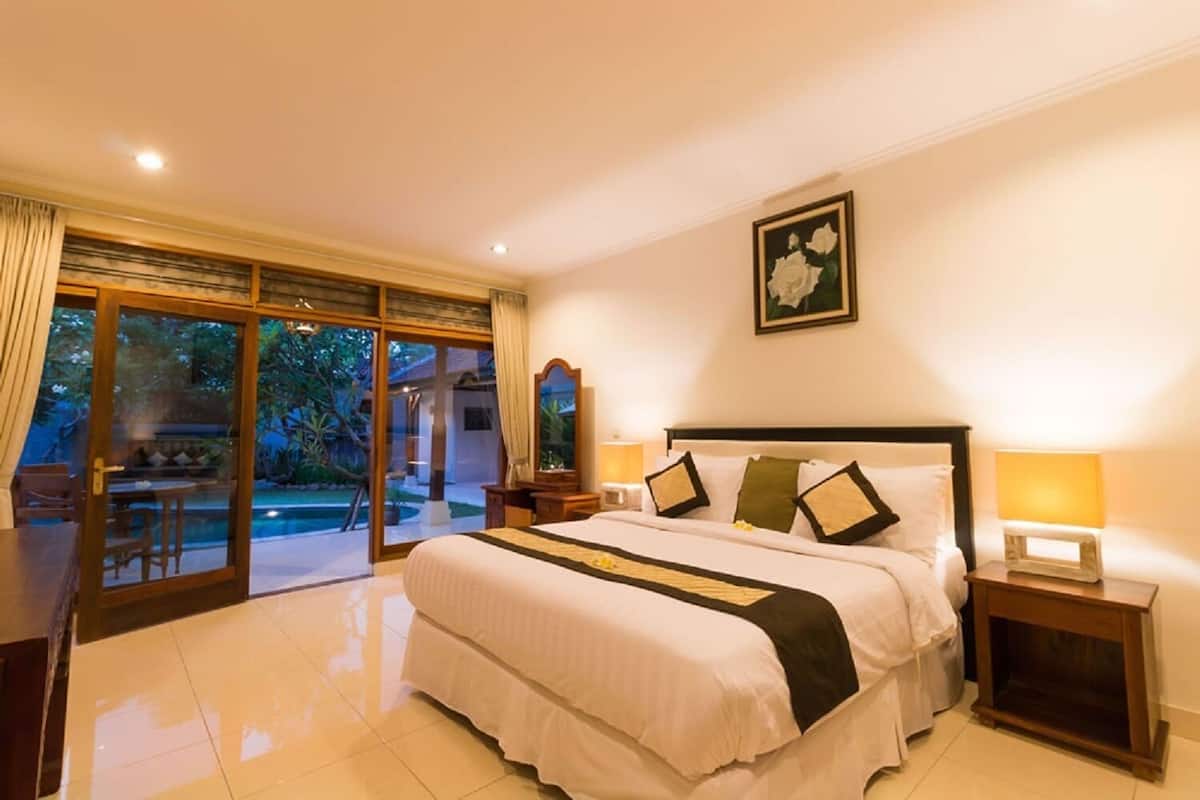 Une chambre spacieuse avec un grand lit, une table de chevet, une lampe de chevet et une vue sur une piscine et un jardin, visible à travers des portes vitrées.