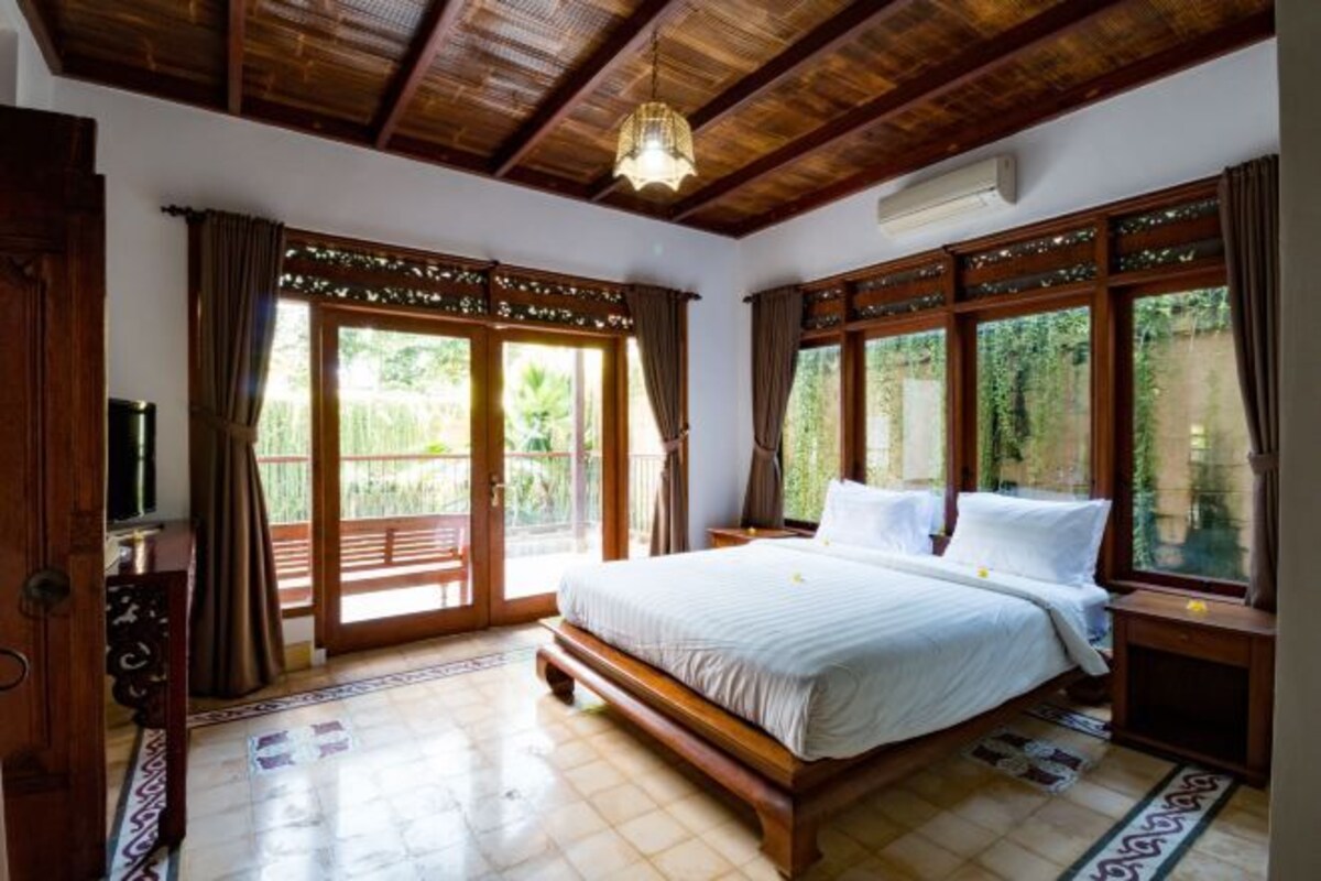 Een slaapkamer met een groot bed, houten plafond en grote ramen voorzien van gordijnen.