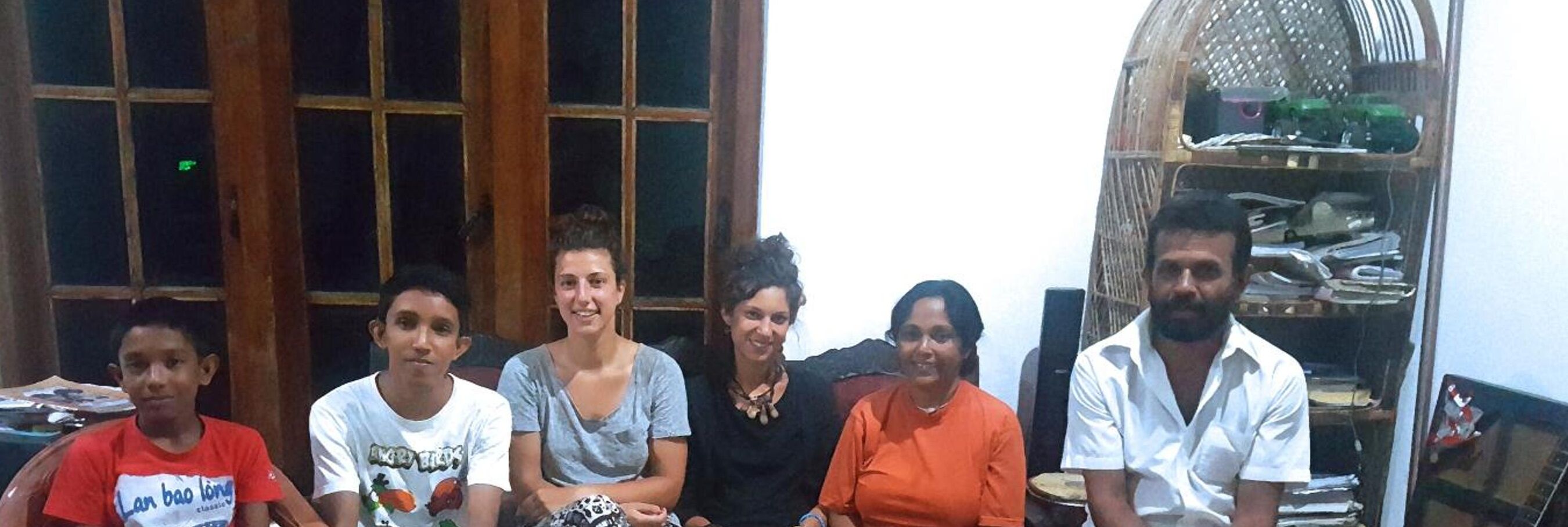 A group of people seated in a room with a wooden window frame and a shelf with various items.