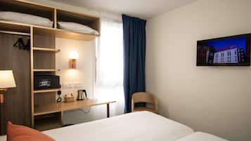 A hotel room with a bed, a desk with a lamp, a chair, a TV on the wall, and a window with curtains.