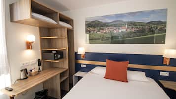 A hotel room with a bed, a desk, a chair, a trash bin, a lamp, a phone, a basket, and a painting of a village on the wall.