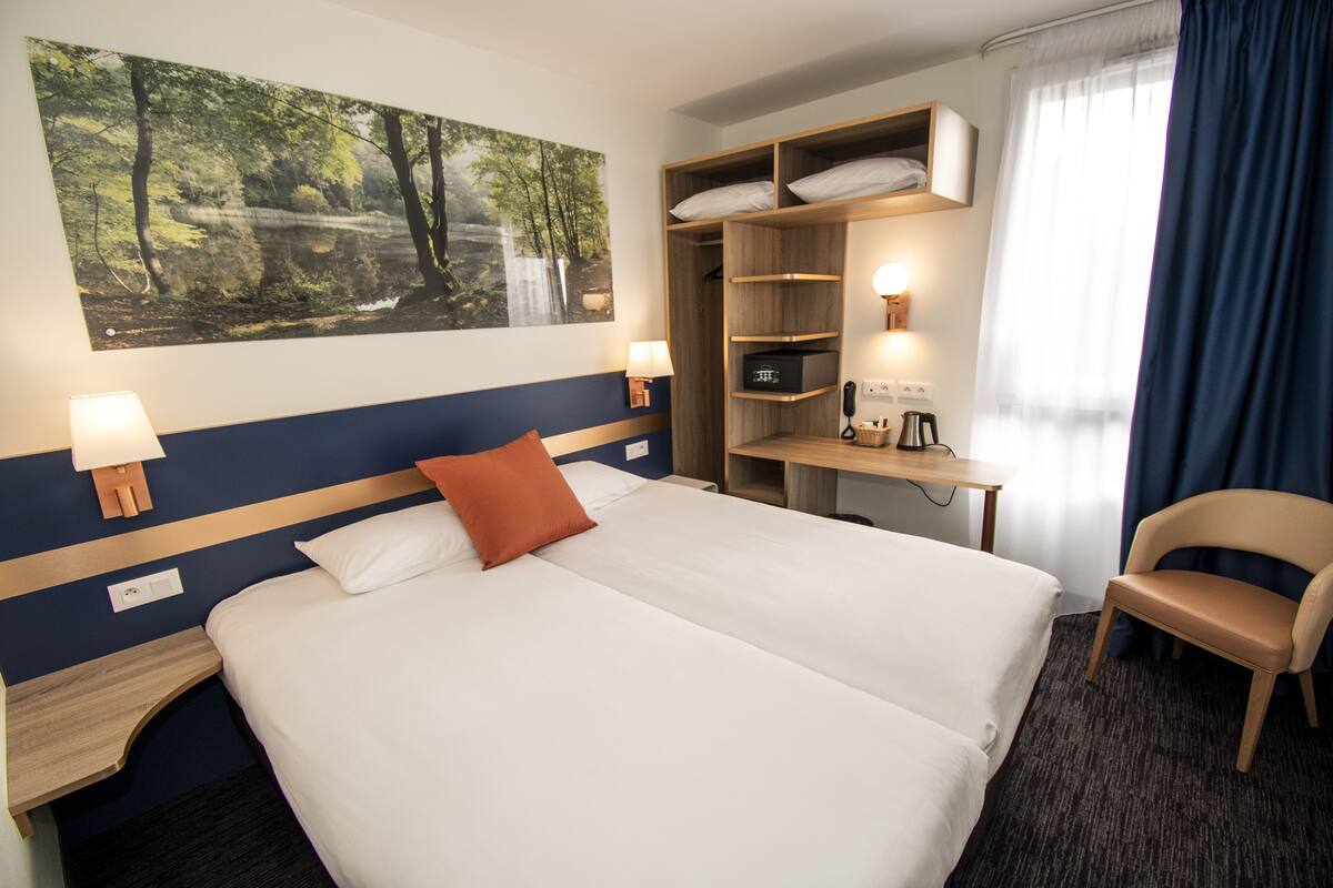 A hotel room with a large bed, a desk with a computer, a chair, a wardrobe, and a painting of a forest on the wall.