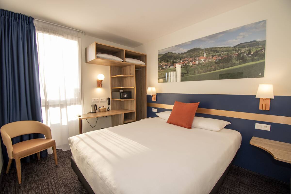 A hotel room with a large bed, a chair, a desk with a lamp, a shelf with a basket, and a painting of a landscape on the wall.