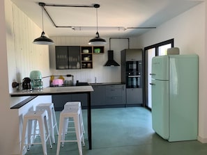 Cozinha compacta com design moderno, com forno embutido, geladeira e pia. Há dois taburetes brancos e um pequeno balcão com um eletrodoméstico rosa.