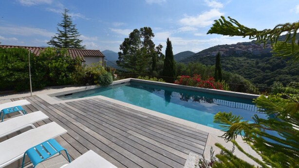 A rectangular swimming pool with a wooden deck, surrounded by greenery and trees, with a view of hills in the background. - Appart'sHotel Fior Di Ribba (Sartene)