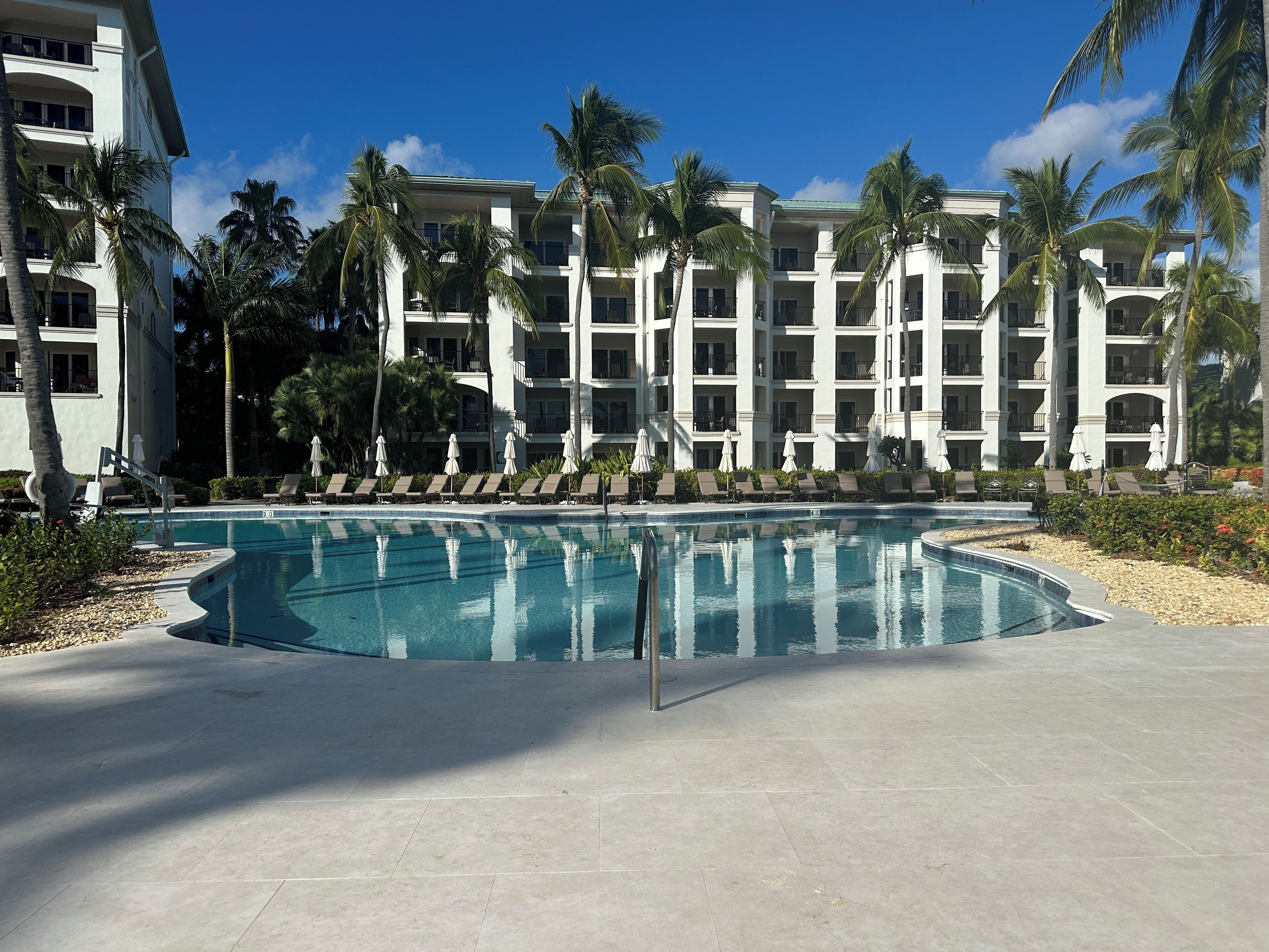 A resort pool with lounge chairs and umbrellas, surrounded by palm trees and a multi-story building.