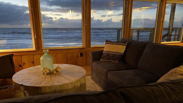 A cozy room with a large window overlooking the ocean, a round wooden table with a decorative bottle, and a dark-colored sofa with patterned cushions. - Oceanfront, Private Beach Access, Newly Updated, Perfect for Family Gatherings! (Lincoln City)