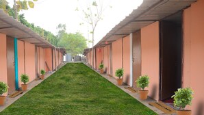 A corridor with a green lawn, potted plants, and doors with a 'Welcome' mat. - Prayag Divine Kumbh Camp - Hostel (Karchana)