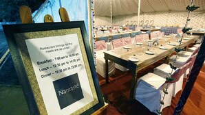 A framed menu board with restaurant timings and a tented dining area with tables set for a meal. - Prayag Divine Kumbh Camp - Hostel (Karchana)